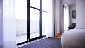 Una habitación de hotel moderna con un amplio ventanal corredero al exterior, una cama con cabecera blanca, una alfombra gris oscuro y una sola silla.