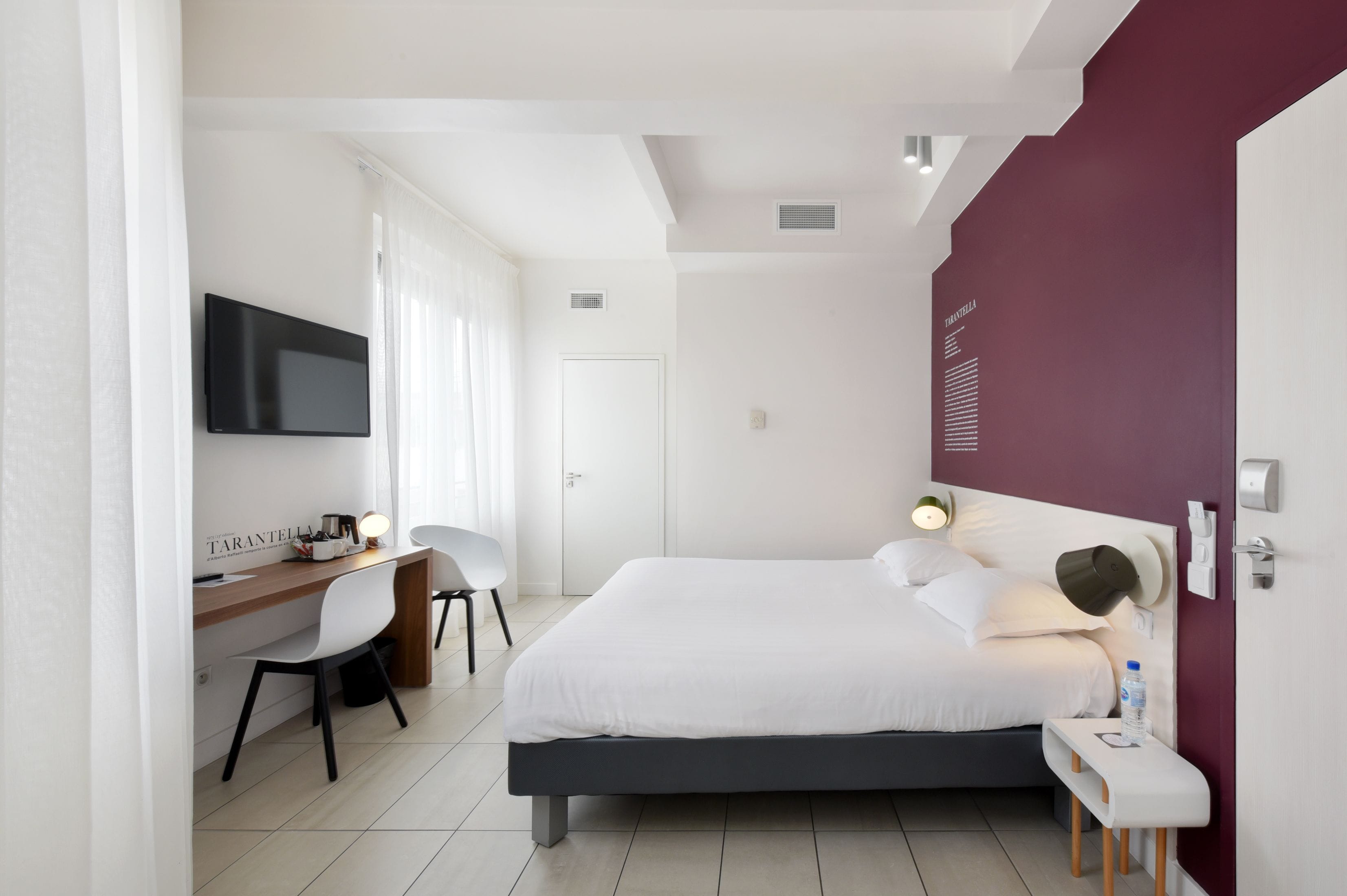 A modern hotel room with a large bed, a desk with a chair, a TV, and a red accent wall.