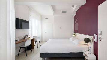 A modern hotel room with a large bed, a desk with a chair, a TV, and a red accent wall.