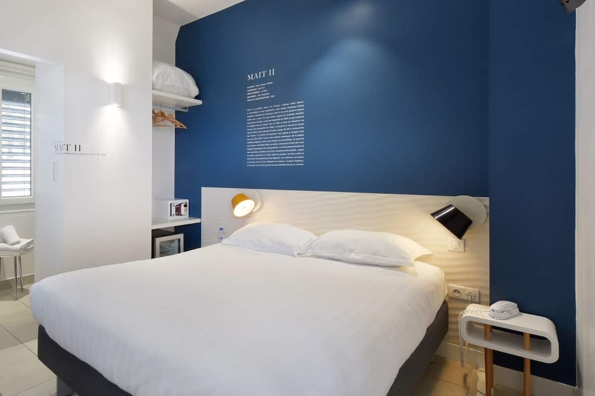 Ein modernes Hotelzimmer mit einem großen Bett, einem Nachttisch mit Lampe und einer an der Wand befestigten Infotafel.