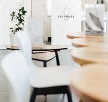 Un área de recepción de hotel moderna con un diseño minimalista, que incluye un mostrador de recepción con el nombre del hotel 'LES VOILES' y un logotipo. Hay varias sillas y mesas dispuestas para los huéspedes, y una planta en maceta añade un toque de verdor.