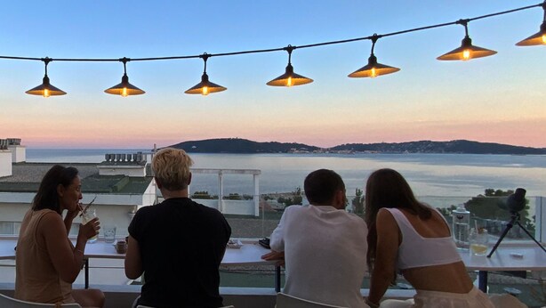 Una terraza con vista al mar, luces colgantes y personas cenando.