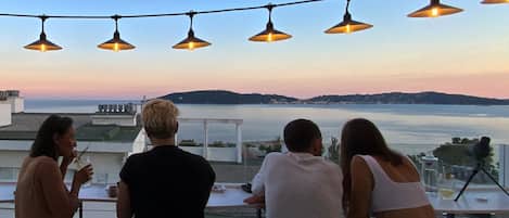 A terrace with a view of the sea, string lights, and people dining.