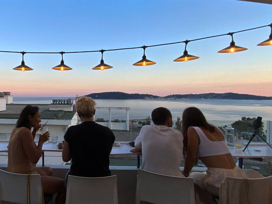 Una terraza con vista al mar, luces colgantes y personas cenando.