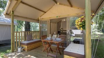 Una terraza de madera con una mesa y bancos, un espacio de descanso techado y vistas al interior.