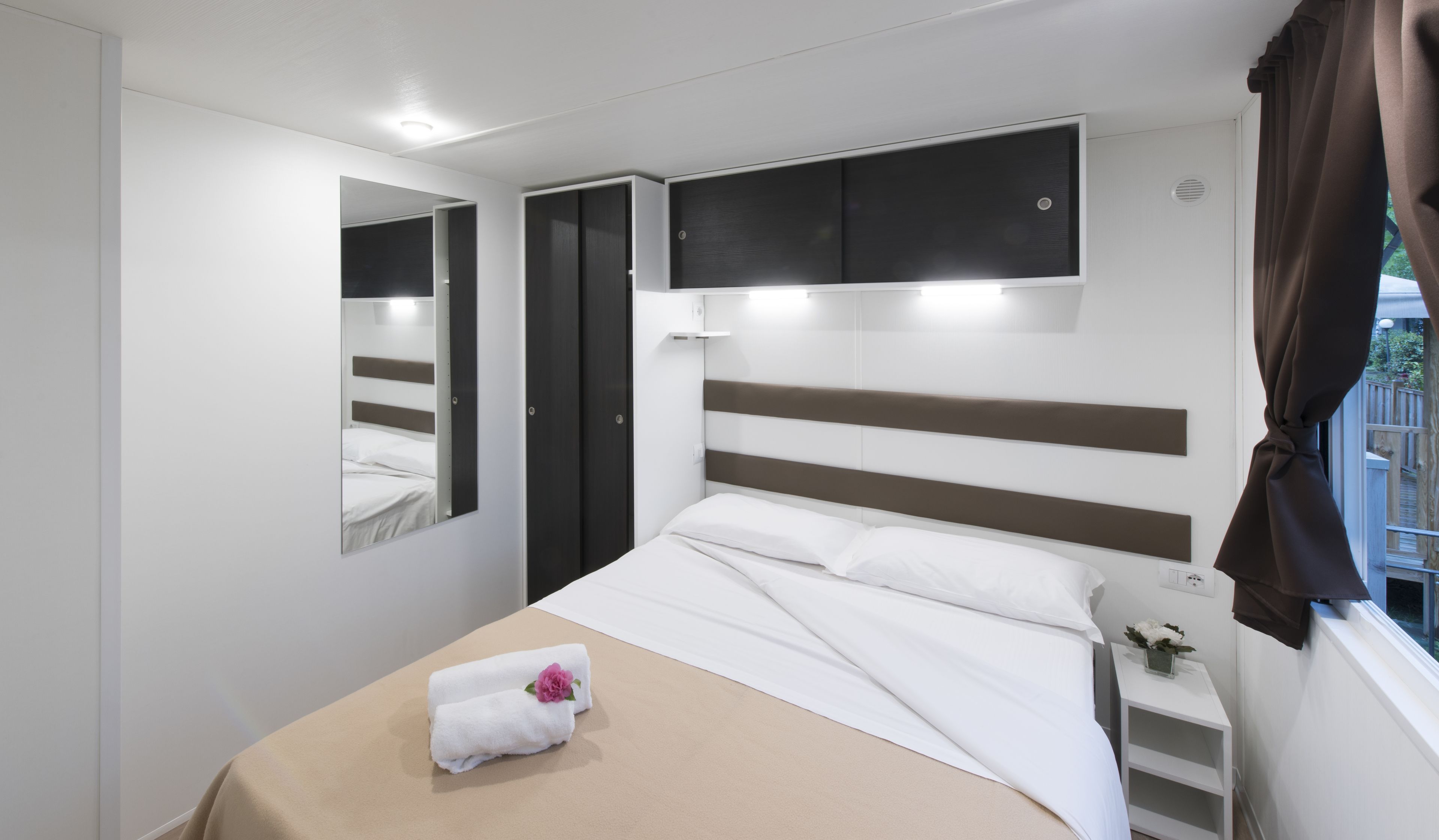 A modern bedroom with a bed, a bedside table, a mirror, and a window with curtains.