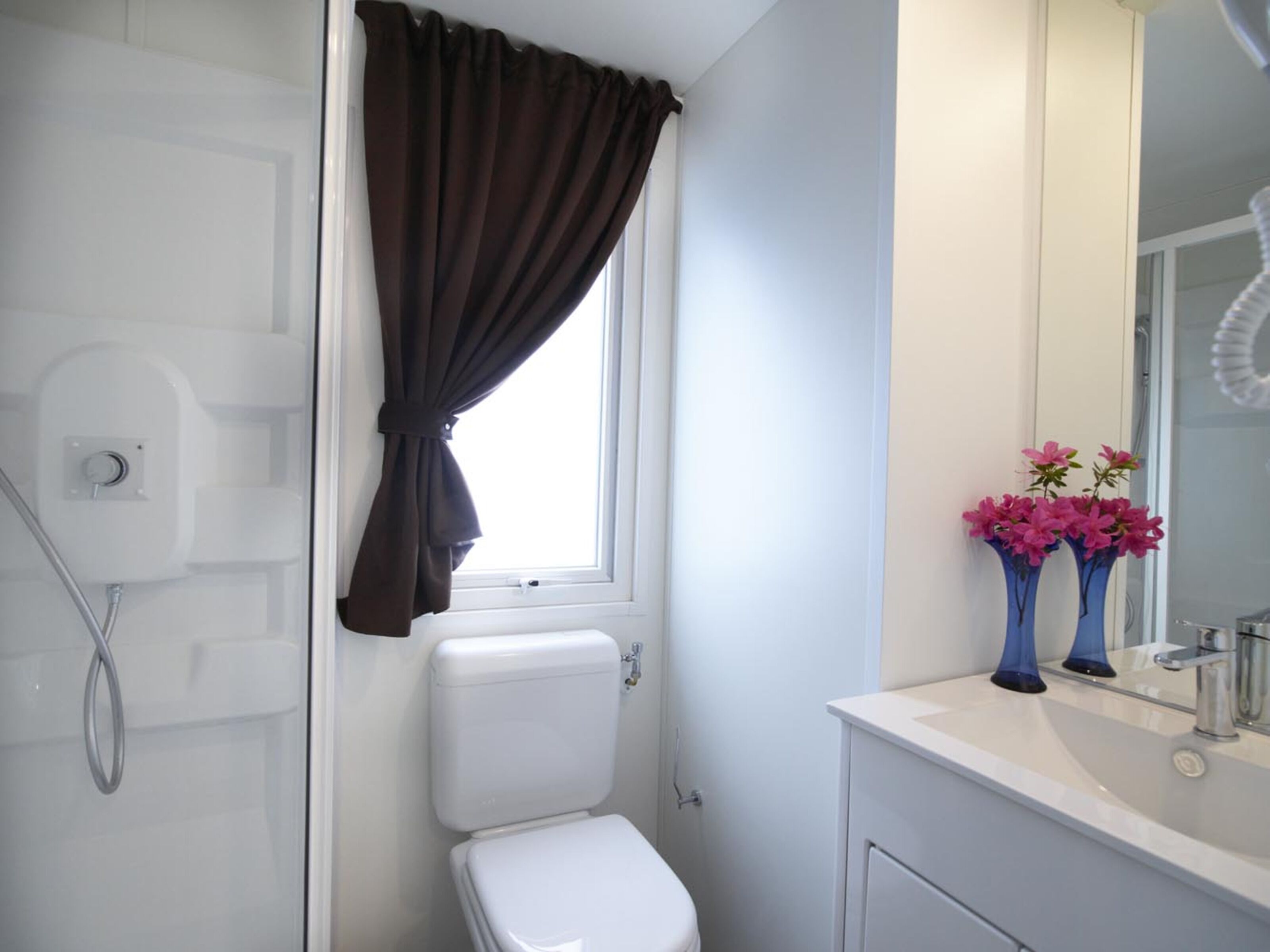 Ein Badezimmer mit Toilette, Dusche und Waschbecken mit Spiegel. Auf dem Waschbecken stehen zwei Vasen mit Blumen.