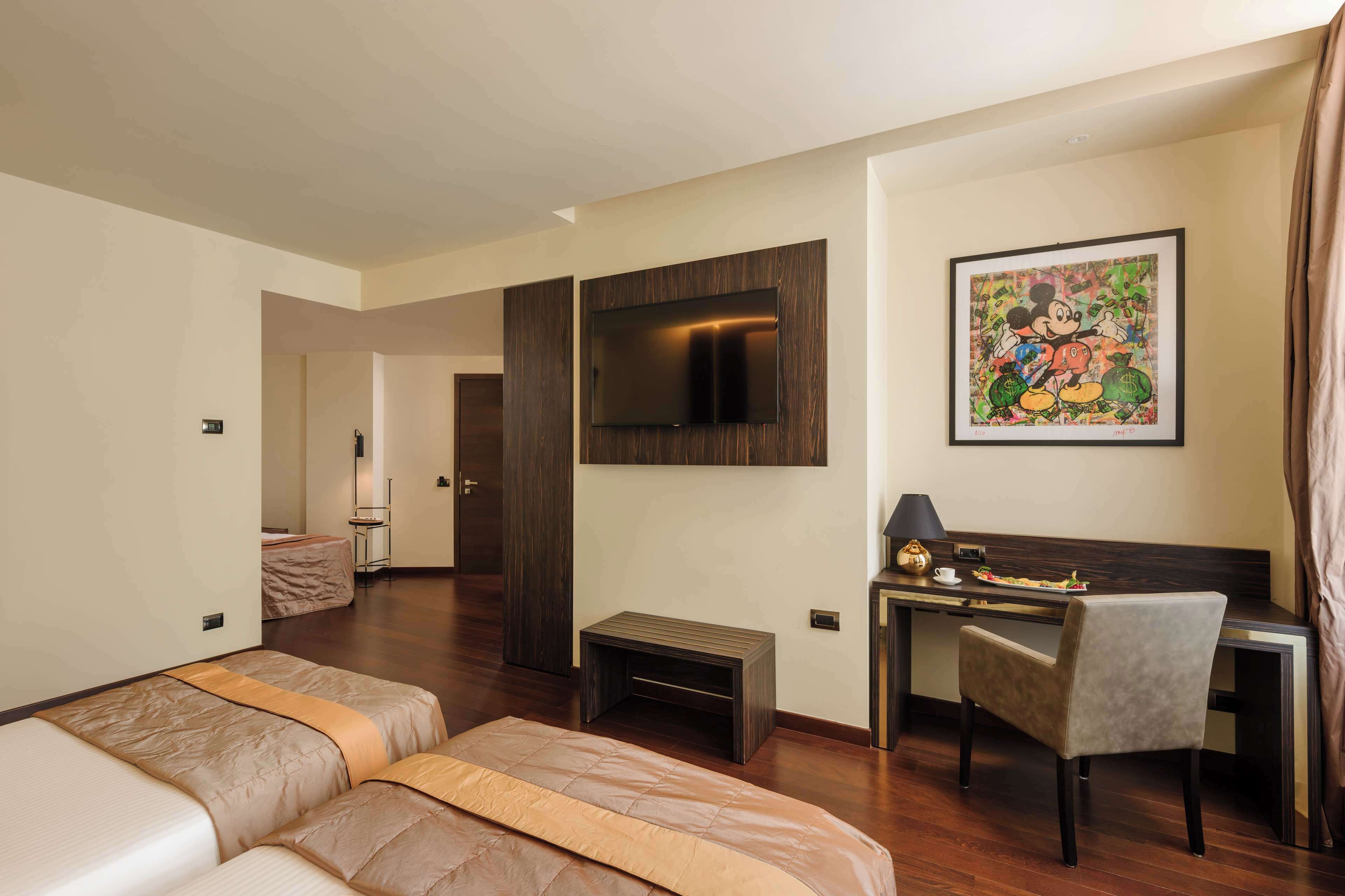 Habitación de hotel con cama, escritorio, silla, televisor montado en la pared y un cuadro enmarcado.