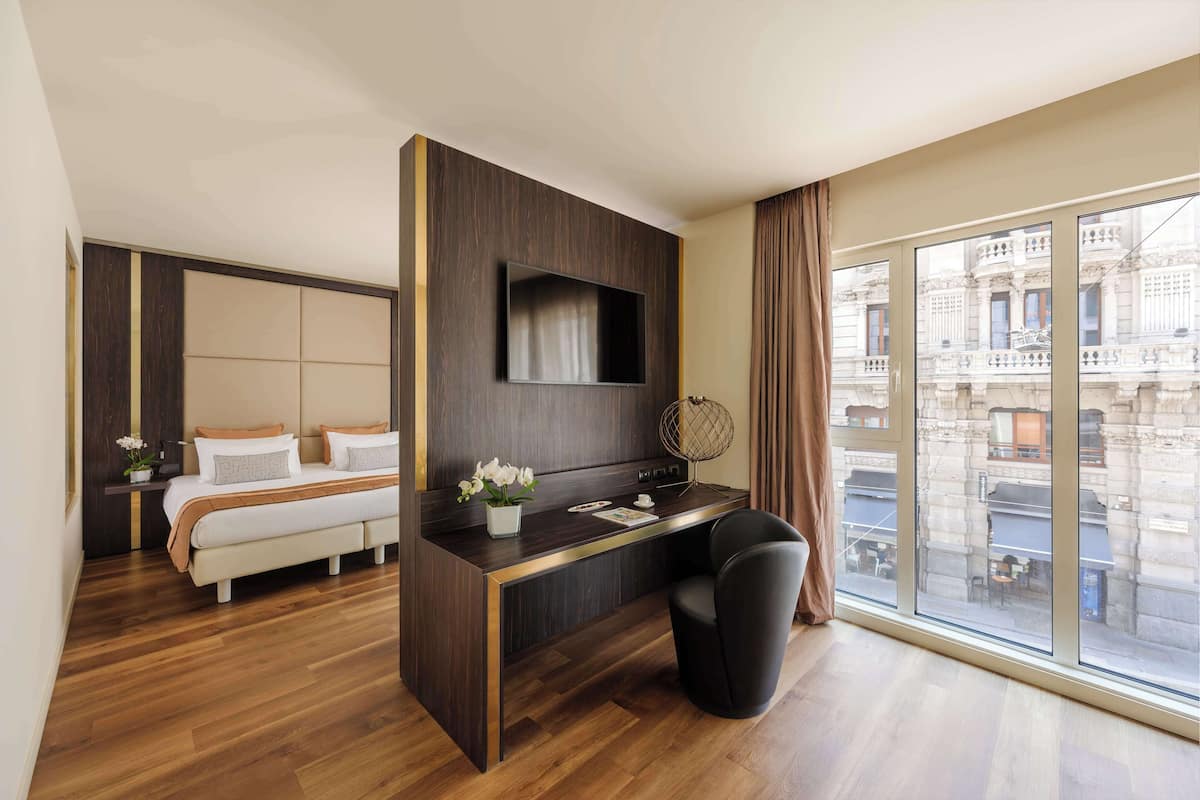 Ett modernt hotellrum med en säng, ett skrivbord, en stol, en TV och stora fönster med utsikt över staden.