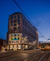 Un hotel de varios pisos con entrada en esquina, tiendas en la planta baja y una línea de tranvía al frente.