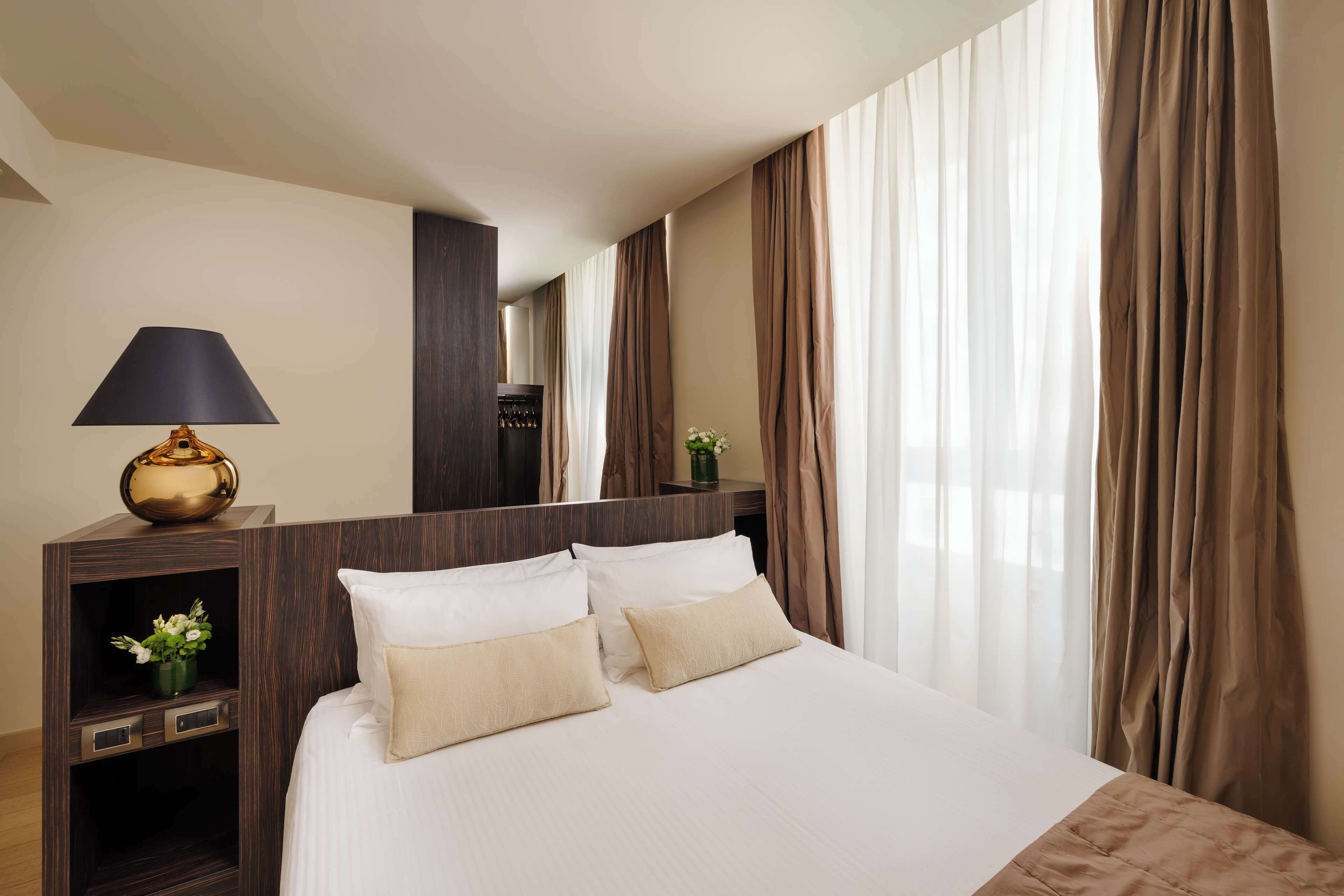 A hotel room with a large bed, a bedside table with a lamp, a mirror, and a view of the outside through sheer curtains.