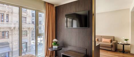 Una habitación de hotel moderna con una cama grande, un televisor de pantalla plana montado en un panel de madera oscura, una mesita redonda con una planta y un sofá con un cojín.