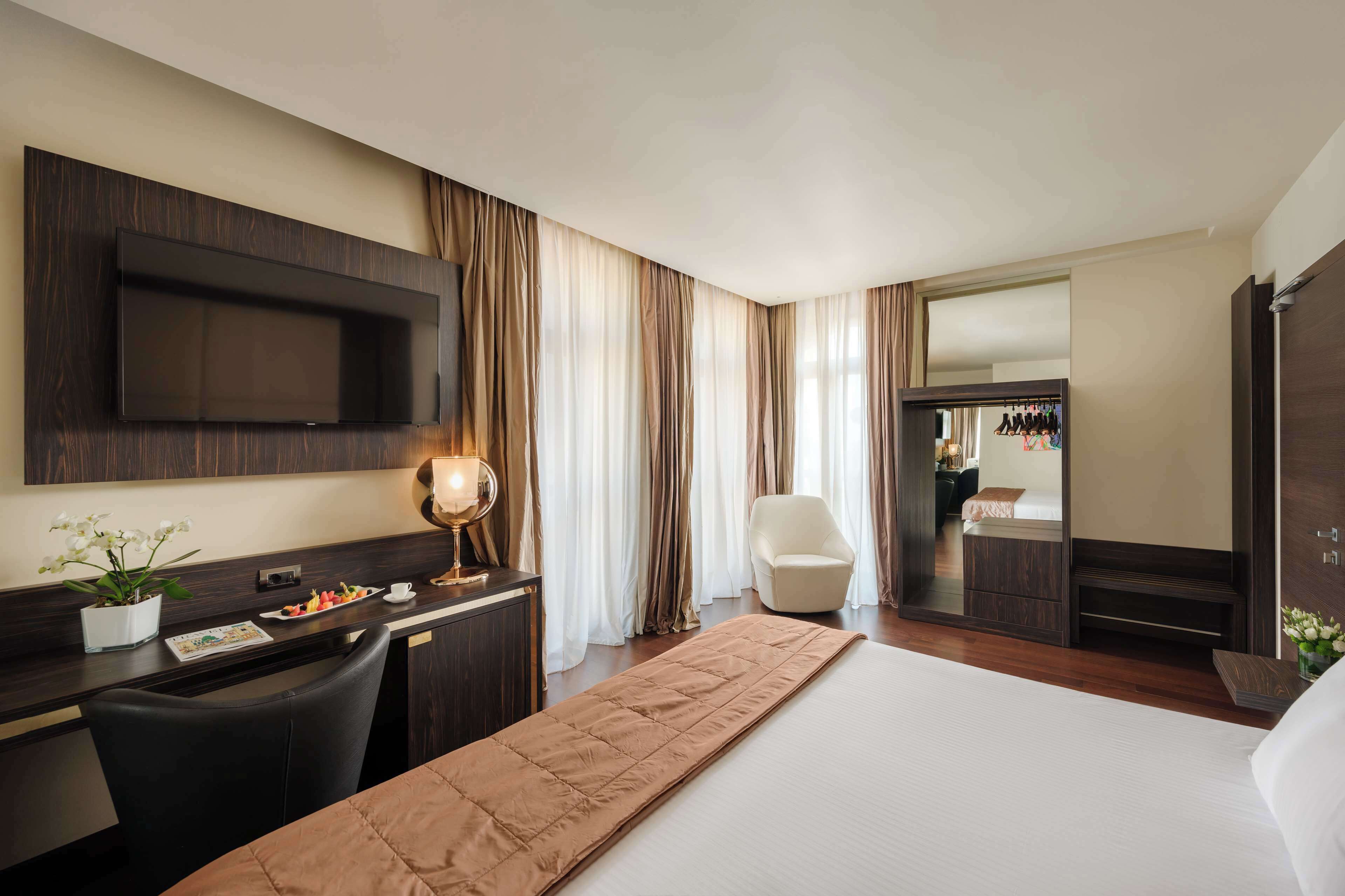 A hotel room with a bed, a desk with a flat-screen TV, a chair, a bedside table with a lamp, and a large mirror.