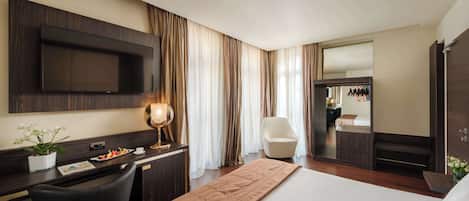 A hotel room with a bed, a desk with a flat-screen TV, a chair, a bedside table with a lamp, and a large mirror.
