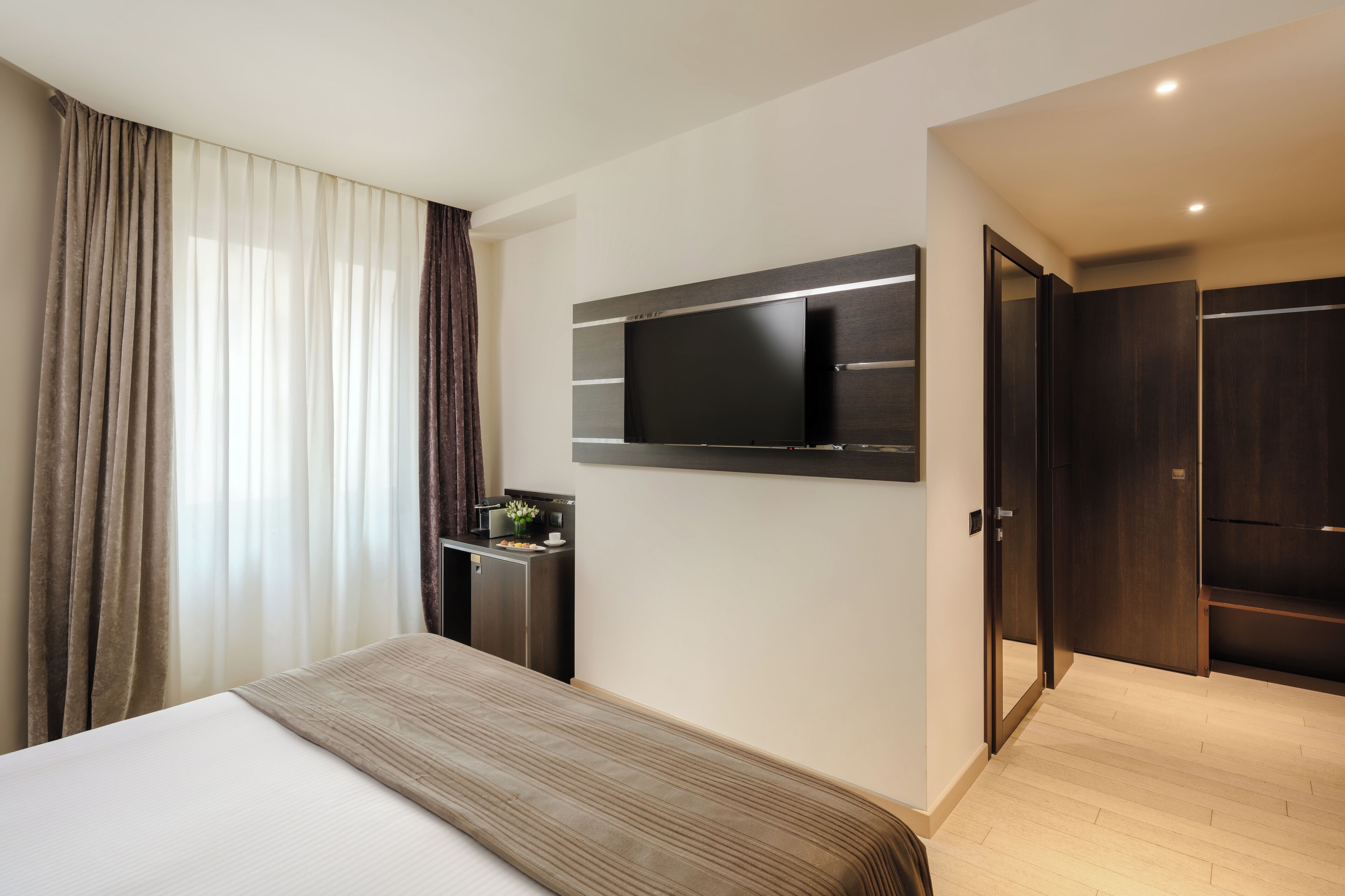 A modern hotel room with a large bed, a flat-screen TV mounted on the wall, a small desk with a lamp, and a wardrobe.
