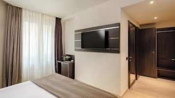 A modern hotel room with a large bed, a flat-screen TV mounted on the wall, a small desk with a lamp, and a wardrobe.