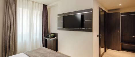 A modern hotel room with a large bed, a flat-screen TV mounted on the wall, a small desk with a lamp, and a wardrobe.