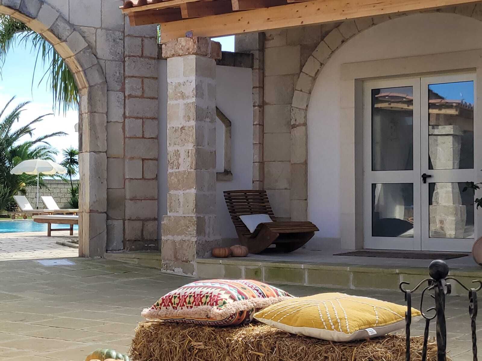 Un área de estar al aire libre con techo, una pared de piedra, un banco de madera y vista a una piscina y palmeras.