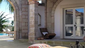 Un área de estar al aire libre con techo, una pared de piedra, un banco de madera y vista a una piscina y palmeras.
