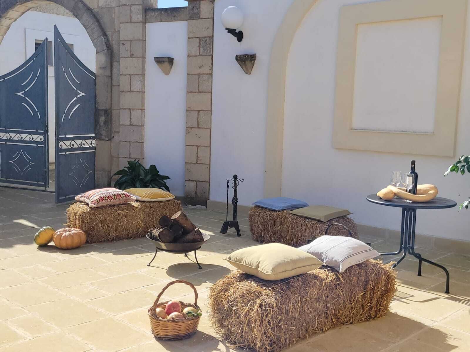 A courtyard with hay bales arranged as seating, a small table with a bottle and glasses, and a basket of fruit.