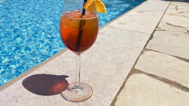Een helderblauw zwembad met ligbedden en parasols, een drankje met een oranje schijfje, en een stenen muur met een bogenpoort op de achtergrond.