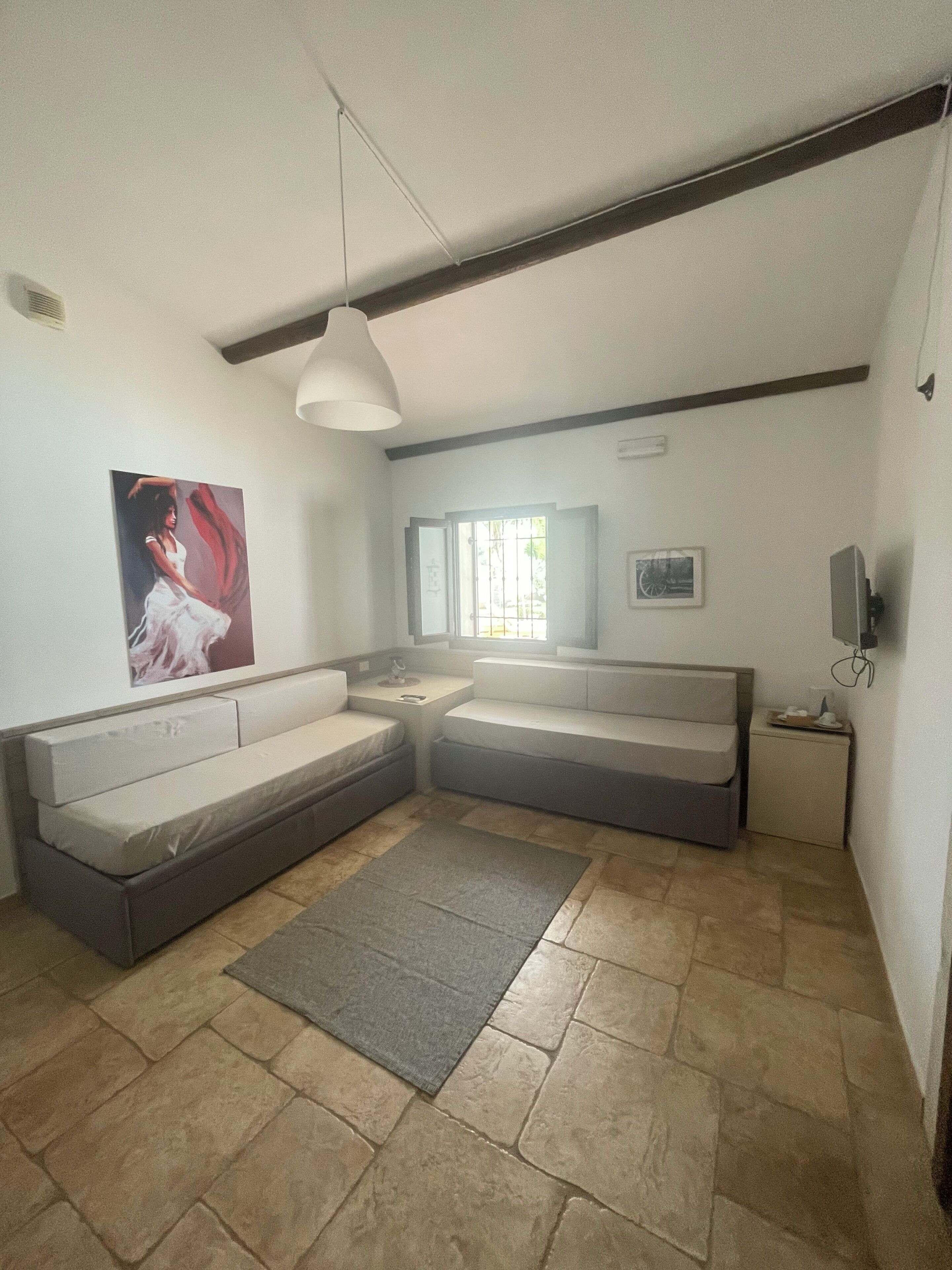 Un salon comprenant un canapé d’angle, un petit tapis, un luminaire au plafond, une fenêtre et un téléviseur fixé au mur.