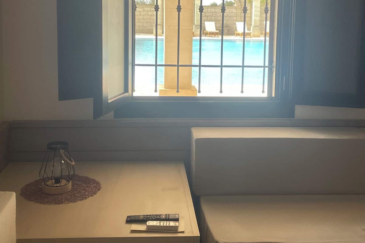 Ein Zimmer mit Blick auf einen Pool durch ein Fenster mit Metallgitter. Auf einem kleinen Tisch liegen ein Fernseher-Sender und eine Kerze.