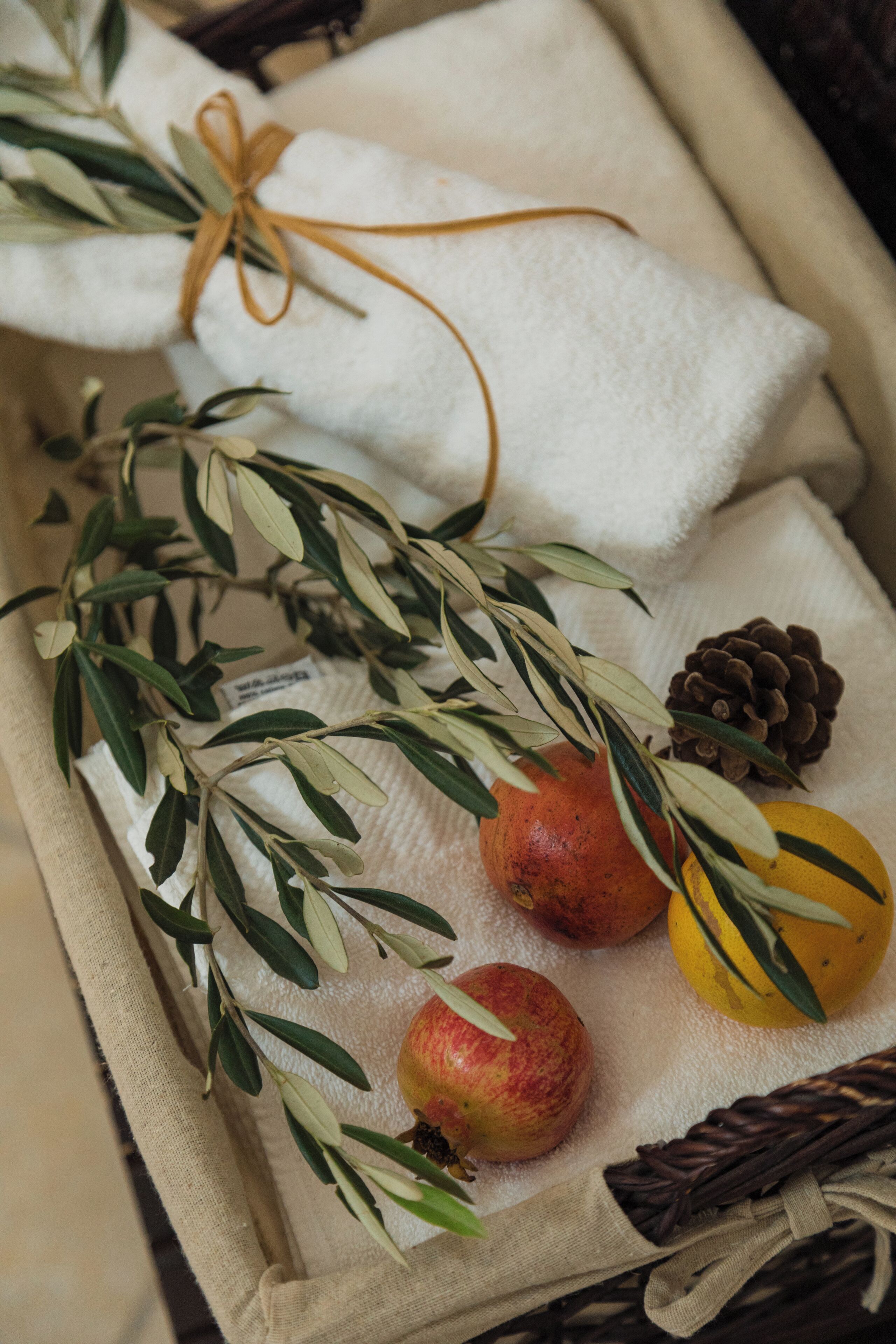 A wicker basket containing a pomegranate, a lemon, a pine cone, and a sprig of olive branches tied with a brown ribbon.