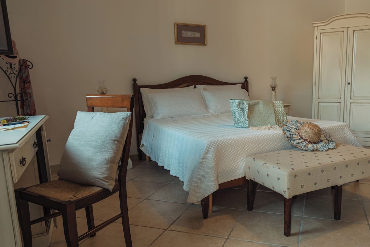 Ein Schlafzimmer mit einem hölzernen Bett, einem Nachttisch, einem Sessel, einem Kleiderschrank und einem kleinen Tisch mit einer Lampe.