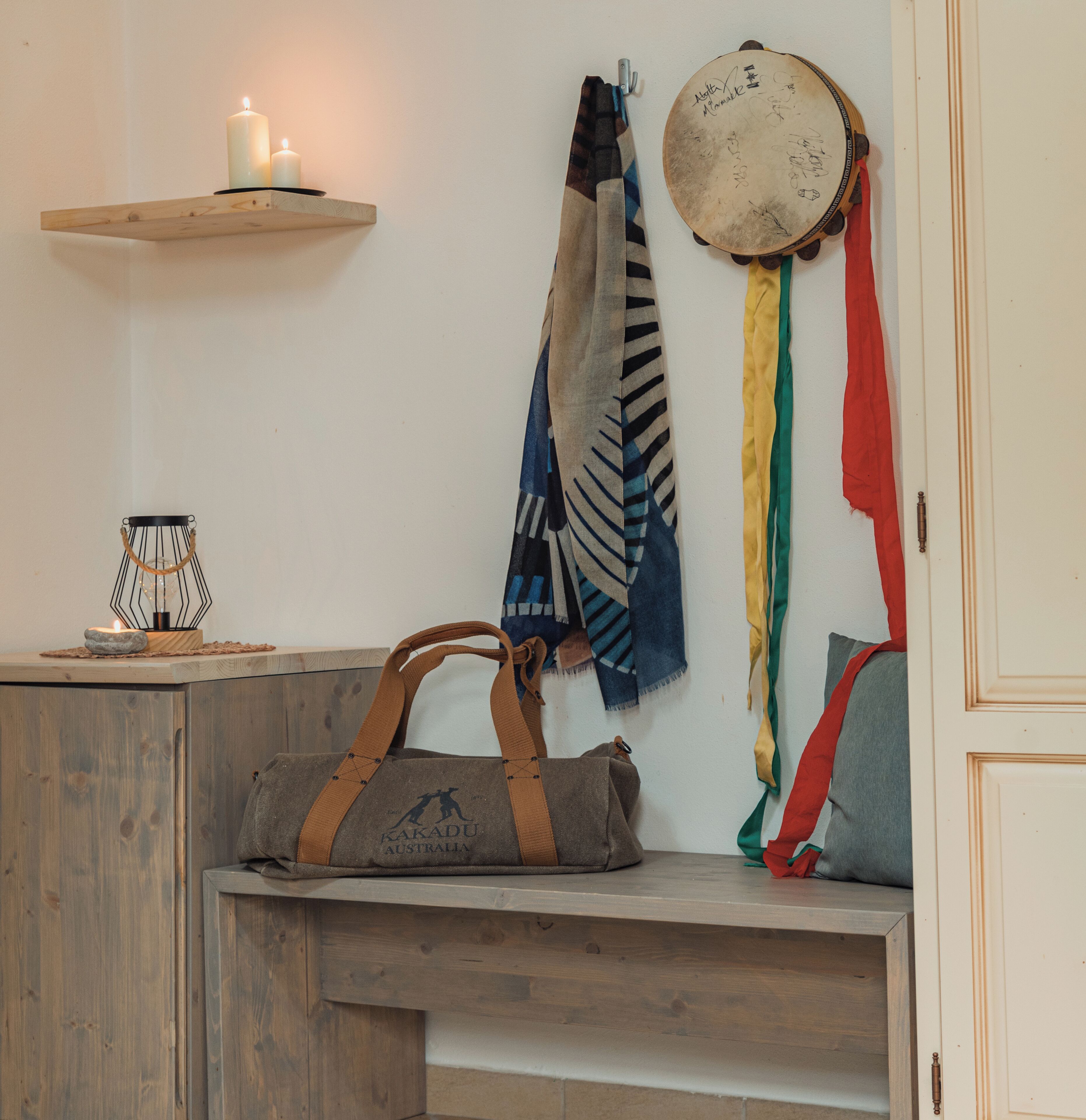 A room with a wooden bench, a wall-mounted shelf with a candle, a drum, and a bag on the bench.