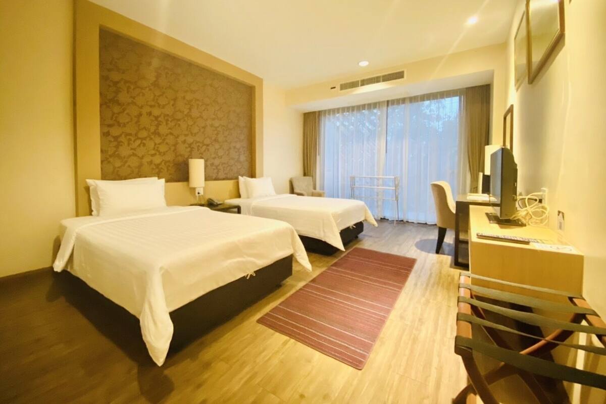 Een hotelkamer met twee bedden, een bureau met een computer, een stoel en een groot raam met gordijnen.