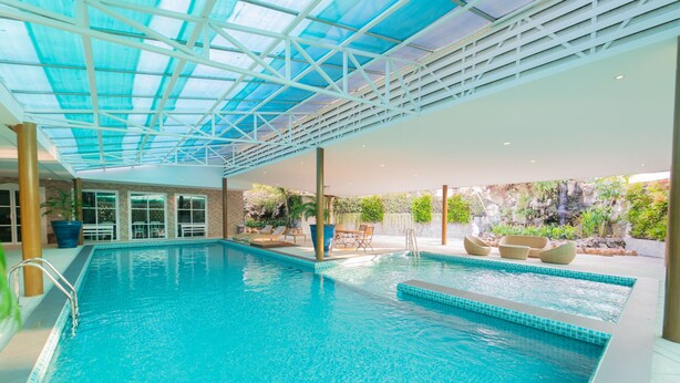 A large indoor swimming pool with a glass ceiling, surrounded by lounge chairs and a waterfall feature. - Rimkhobfa Urban Resort (Samut Prakan)