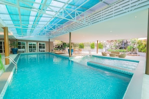 En stor indendørs swimmingpool med glasloft, omgivet af liggestole og et vandfald.