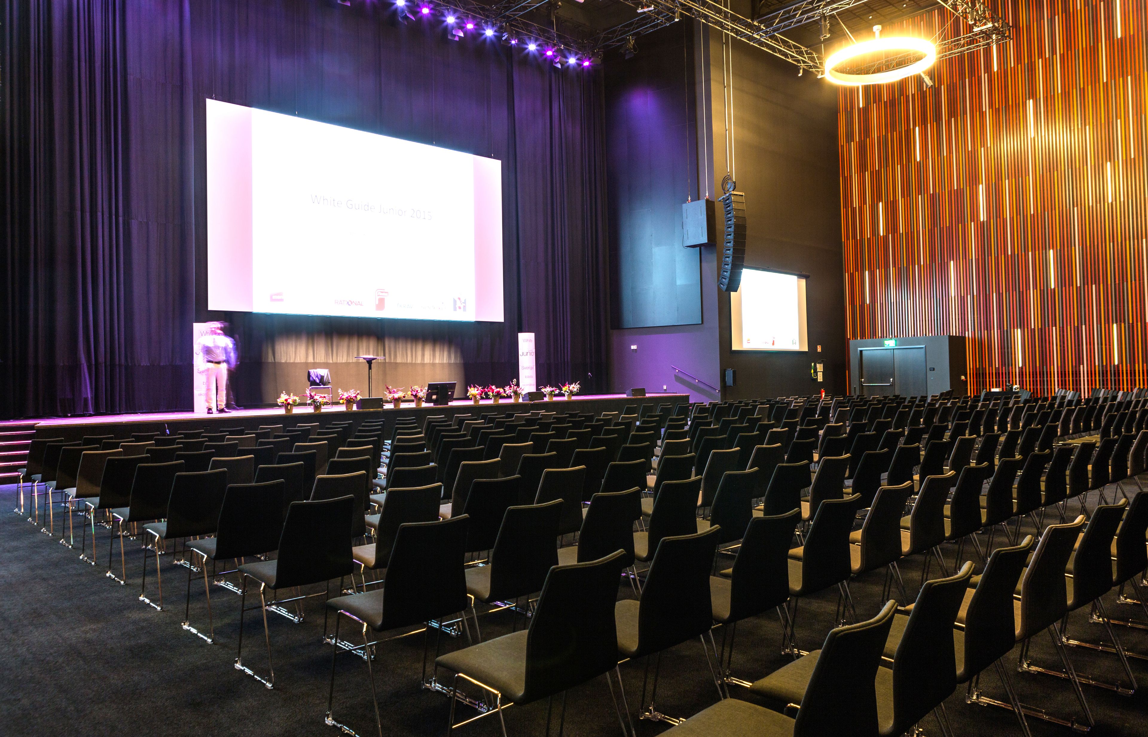 An auditorium with rows of chairs, a stage with a large screen, and a decorative wall.