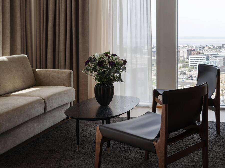 A modern living room with a sofa, a coffee table, and a chair. There is a vase with flowers on the table. The room has a large window with curtains and a view of a cityscape.