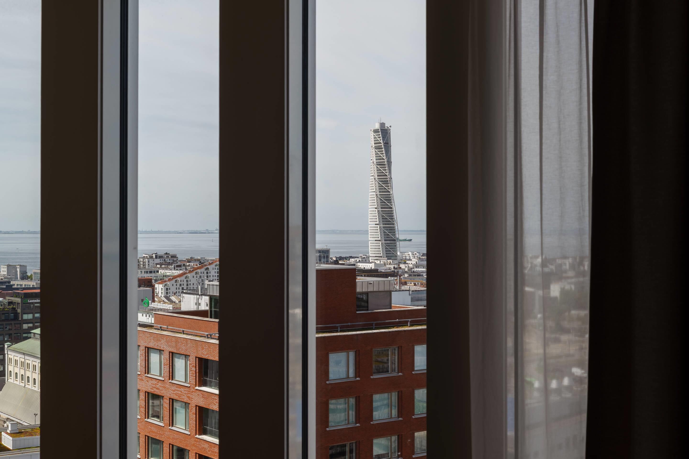 A view through window blinds of a cityscape with a prominent twisting skyscraper in the distance.