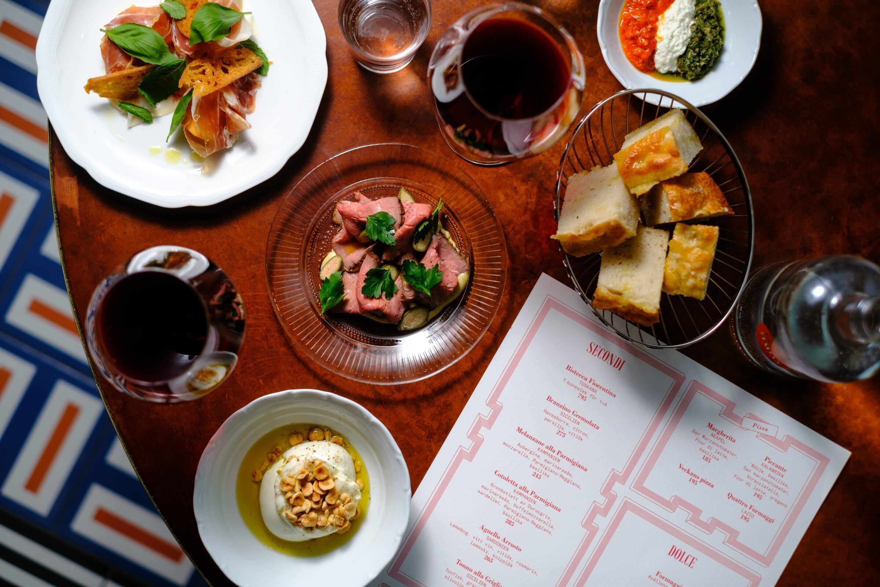 Una mesa con diferentes platos, copas de vino tinto y una carta.