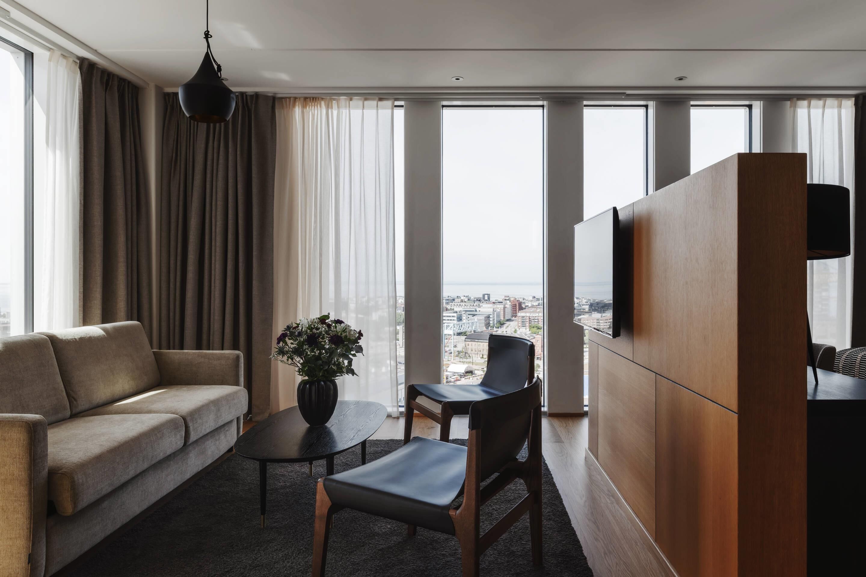 A modern hotel room with a sofa, a small table, a chair, a potted plant, and a large window offering a city view.