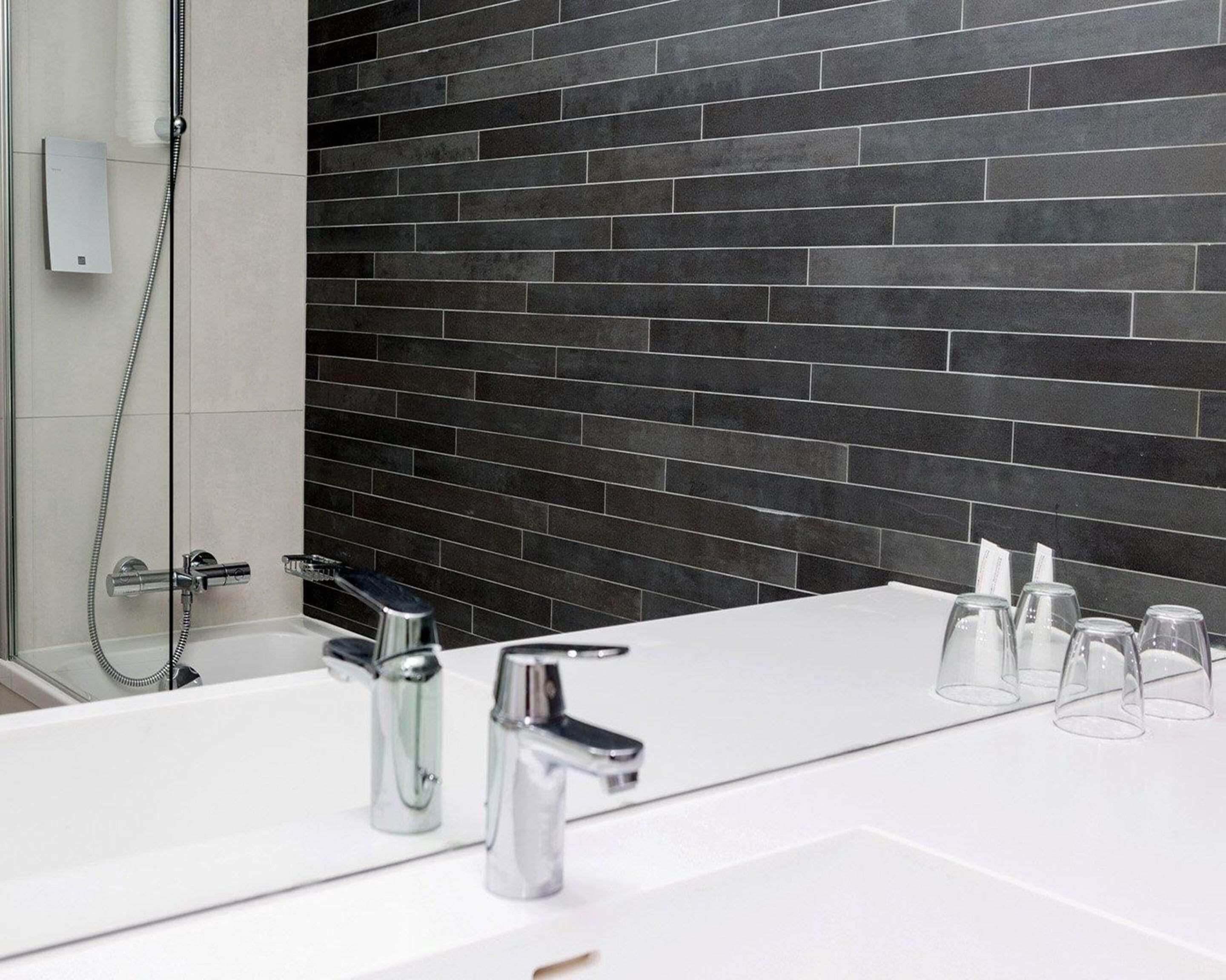 Un baño moderno con un lavabo blanco, dos grifos cromados, un cabezal de ducha de mano y una pared revestida de baldosas oscuras.