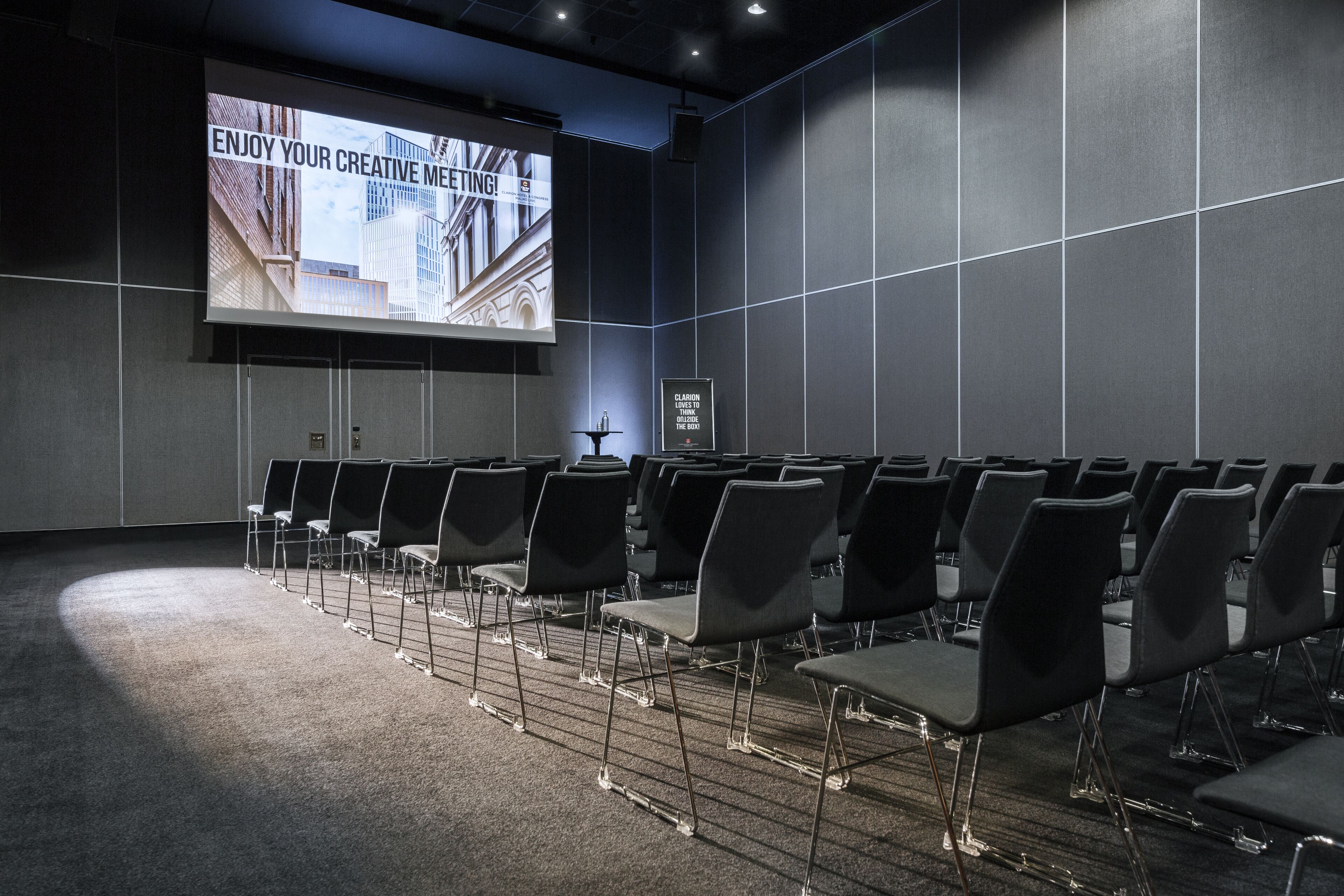 A conference room with rows of chairs, a large screen displaying a presentation, and a framed picture on the wall.