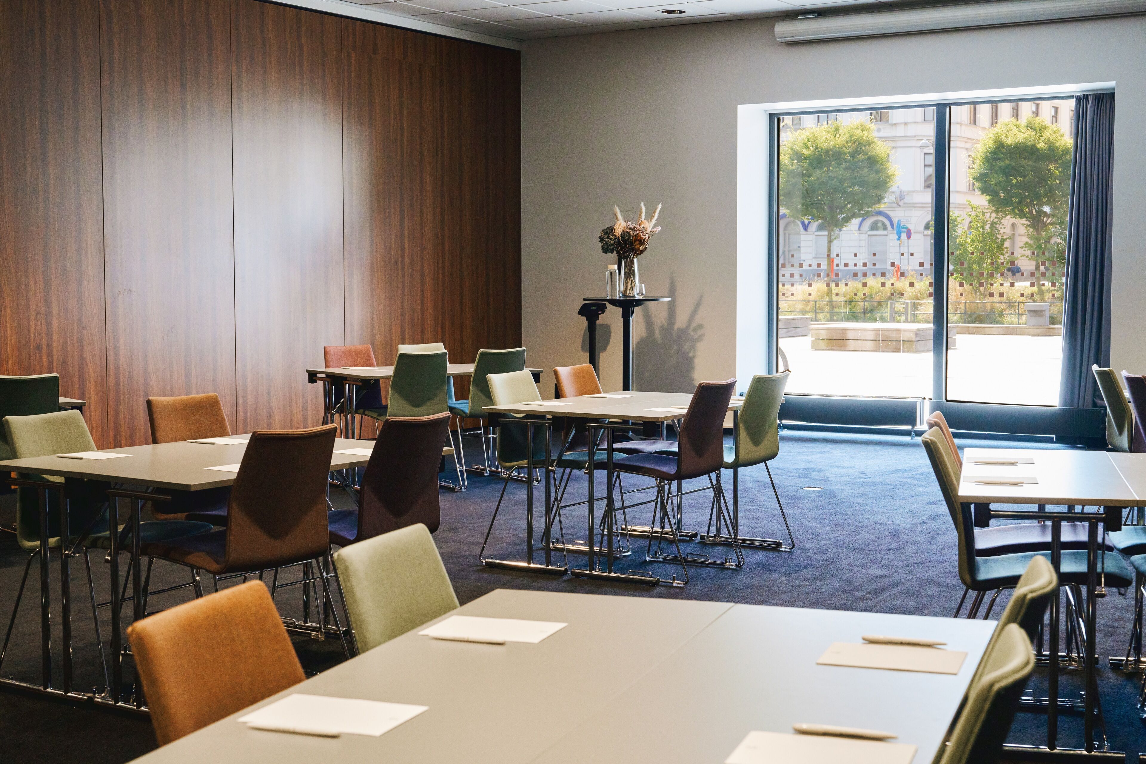 A conference room with multiple tables and chairs, a small table with a vase of dried flowers, and a large window with a view of trees and buildings.