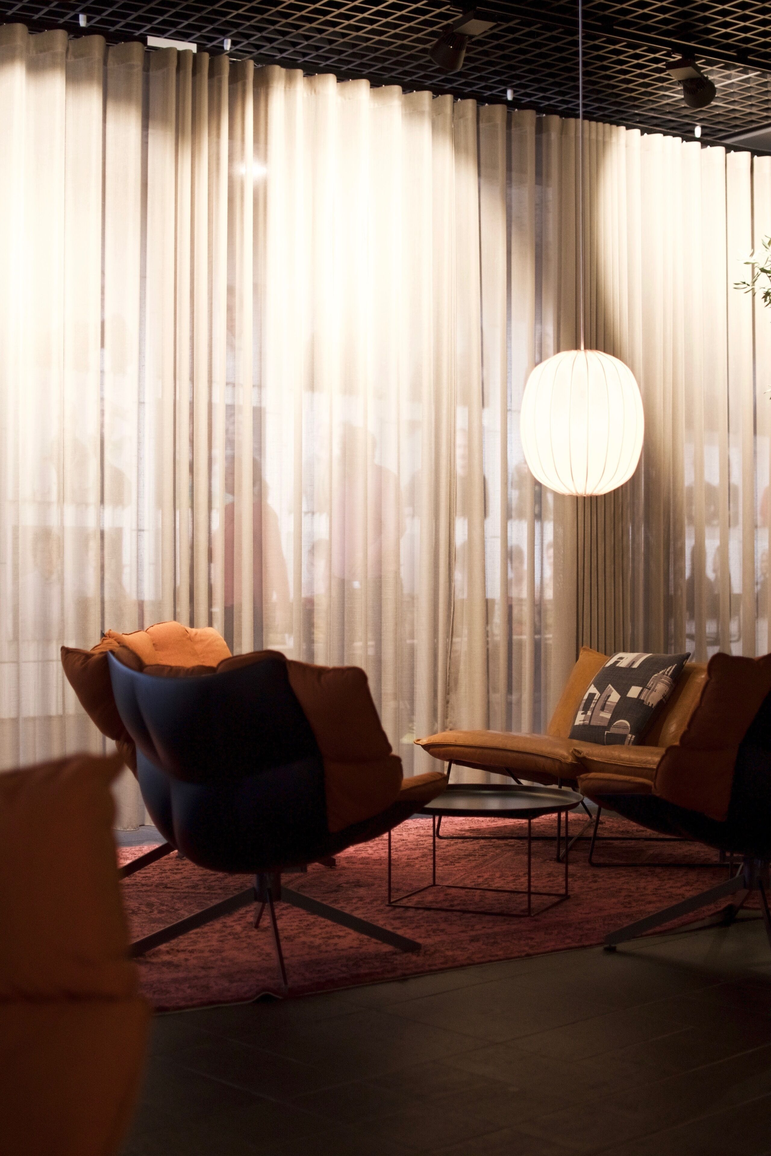 Una habitación con un ventanal grande cubierto por cortinas translúcidas, una mesa redonda, dos sillones y un candelabro colgante.