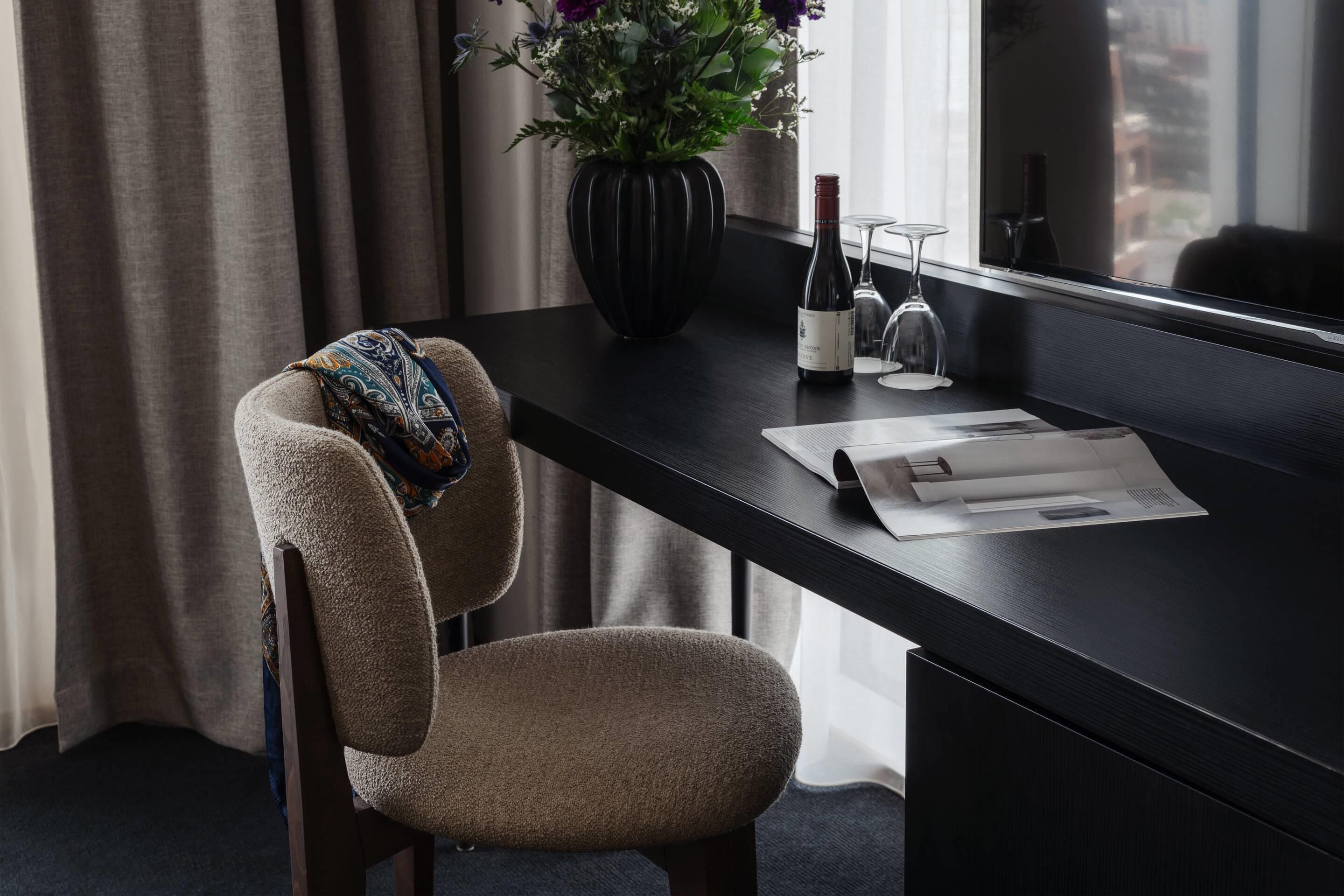 Une chambre d’hôtel avec un bureau en bois foncé, une chaise beige, un vase avec des fleurs, une bouteille de vin et un verre.