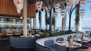 A restaurant with a view of the sea, featuring blue patterned seating, chandeliers, and a modern interior design.