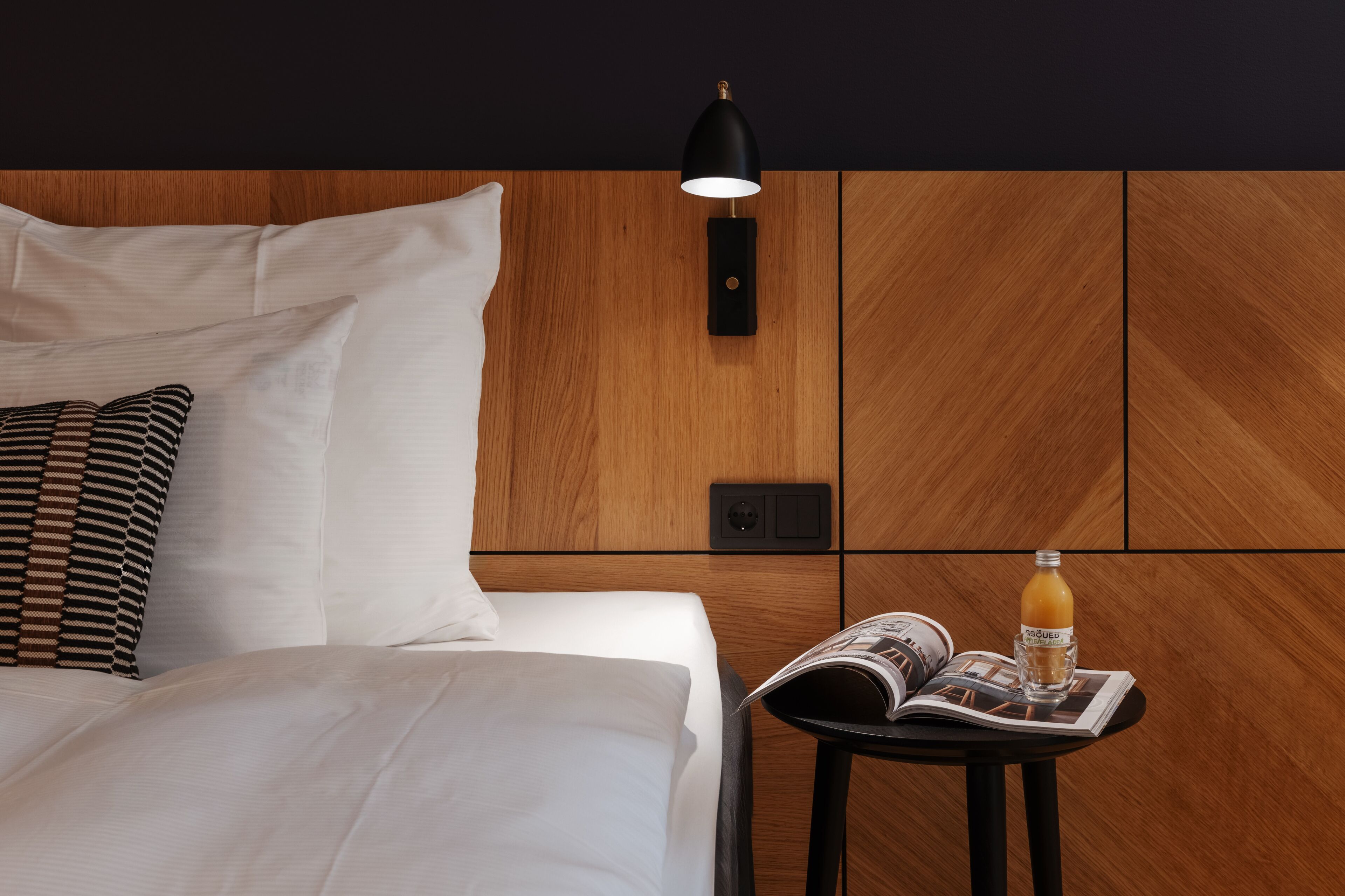Una cama bien hecha con sábanas blancas, una almohada estampada en blanco y negro, una lámpara de noche negra y una mesita con una revista y un vaso de jugo de naranja.