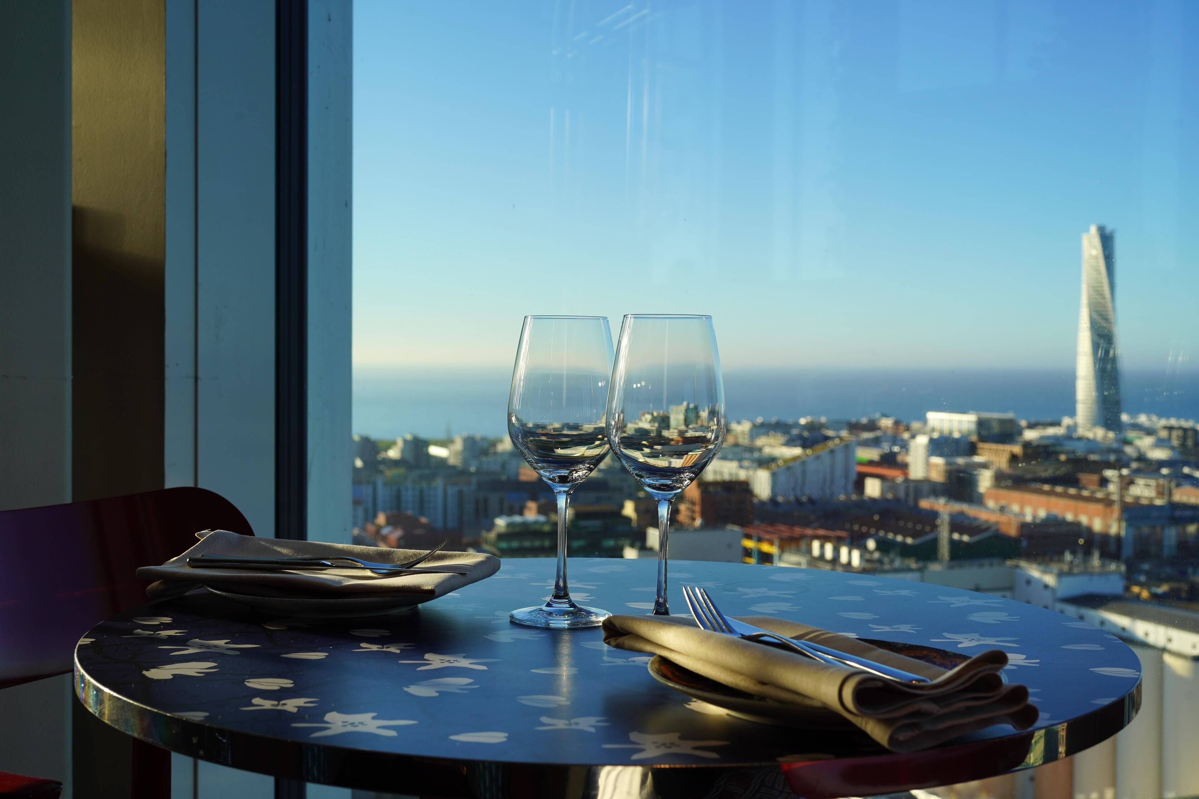 Una mesa con dos copas de vino y un teléfono inteligente, con vista a un paisaje urbano.