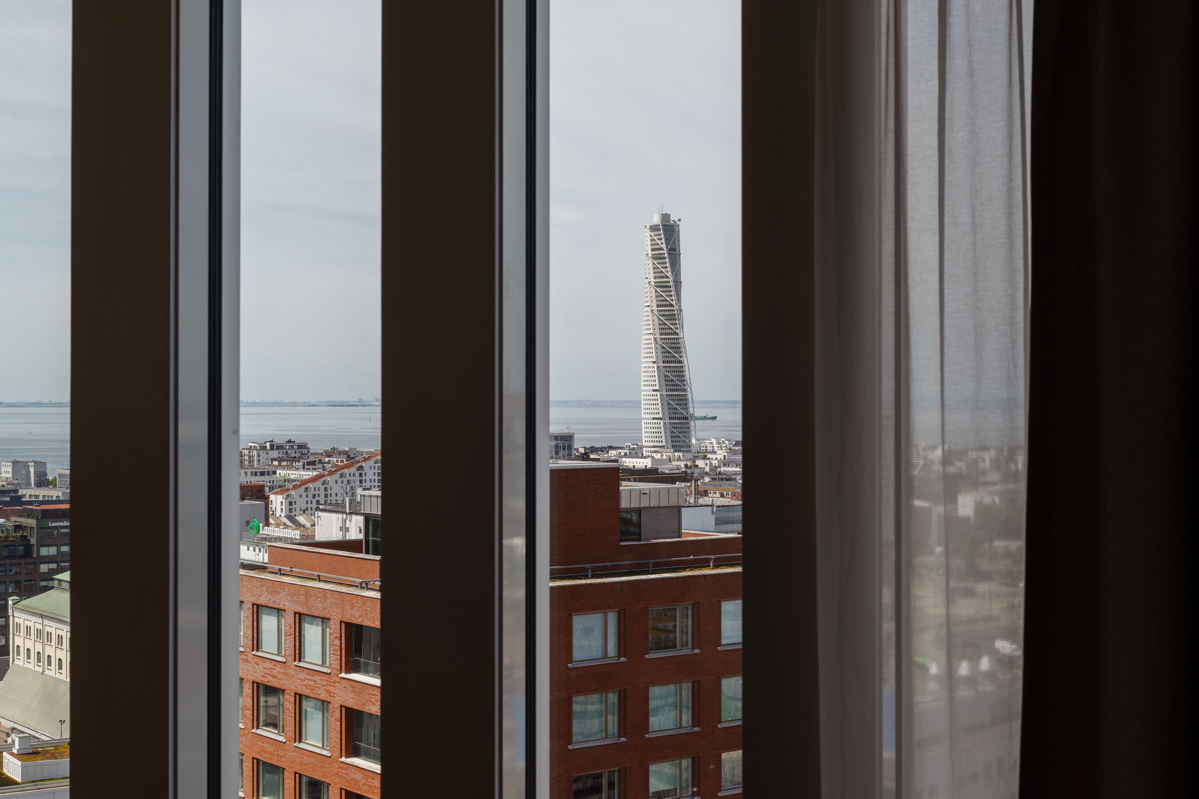 A view through window blinds of a cityscape with a prominent twisting skyscraper and a body of water in the distance.