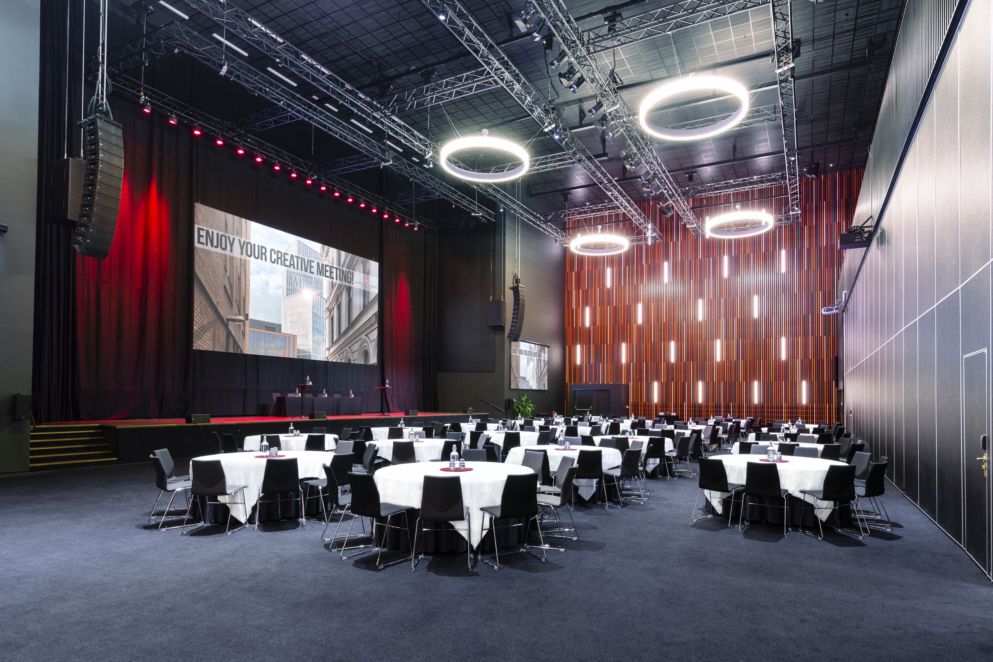 Un salón de conferencias con varias mesas redondas y sillas, un escenario con una pantalla grande e iluminación moderna.
