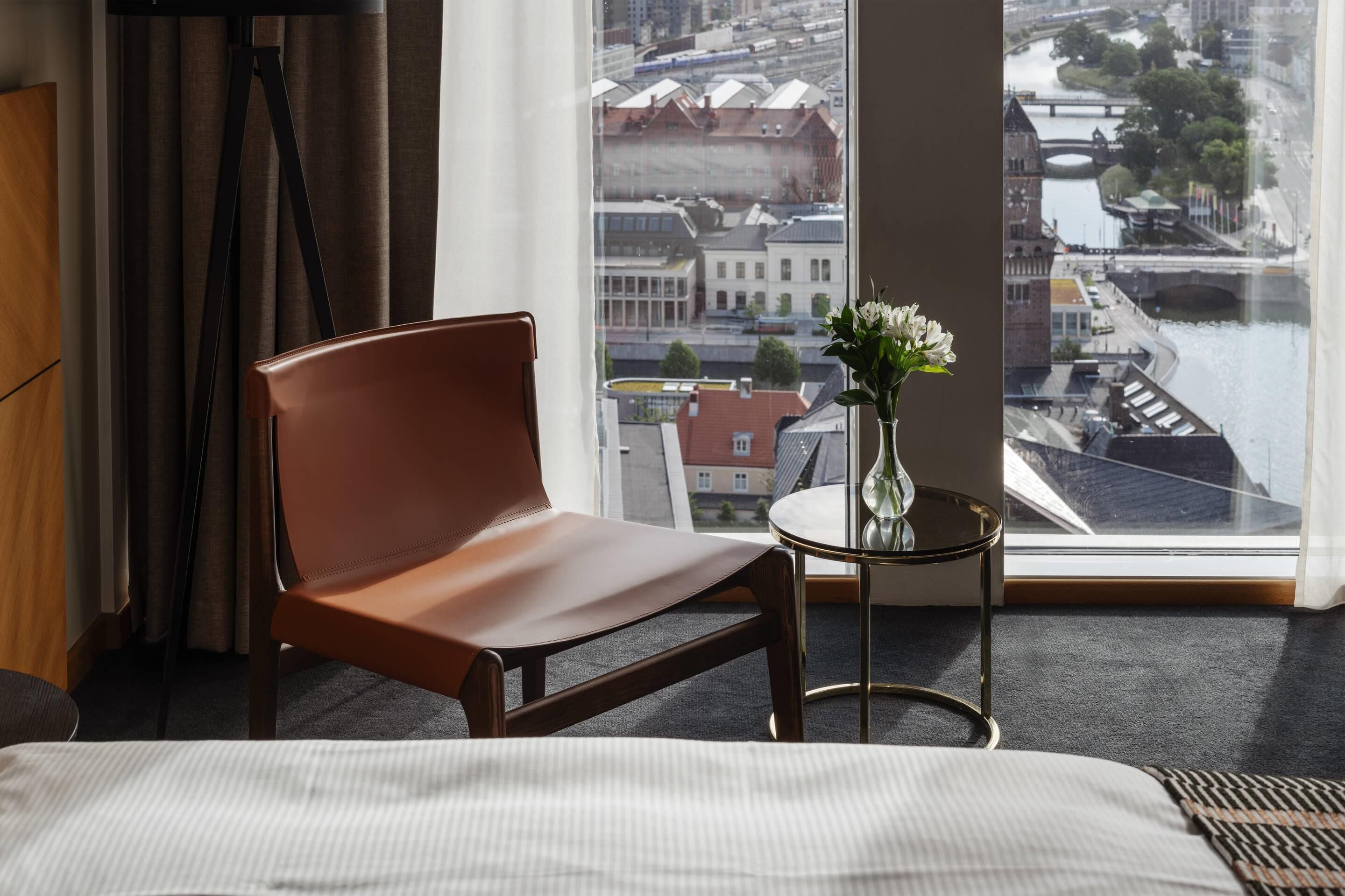 Une chambre d’hôtel avec un lit, un fauteuil en cuir marron, une petite table ronde ornée d’un vase de fleurs et une grande fenêtre donnant sur la ville.