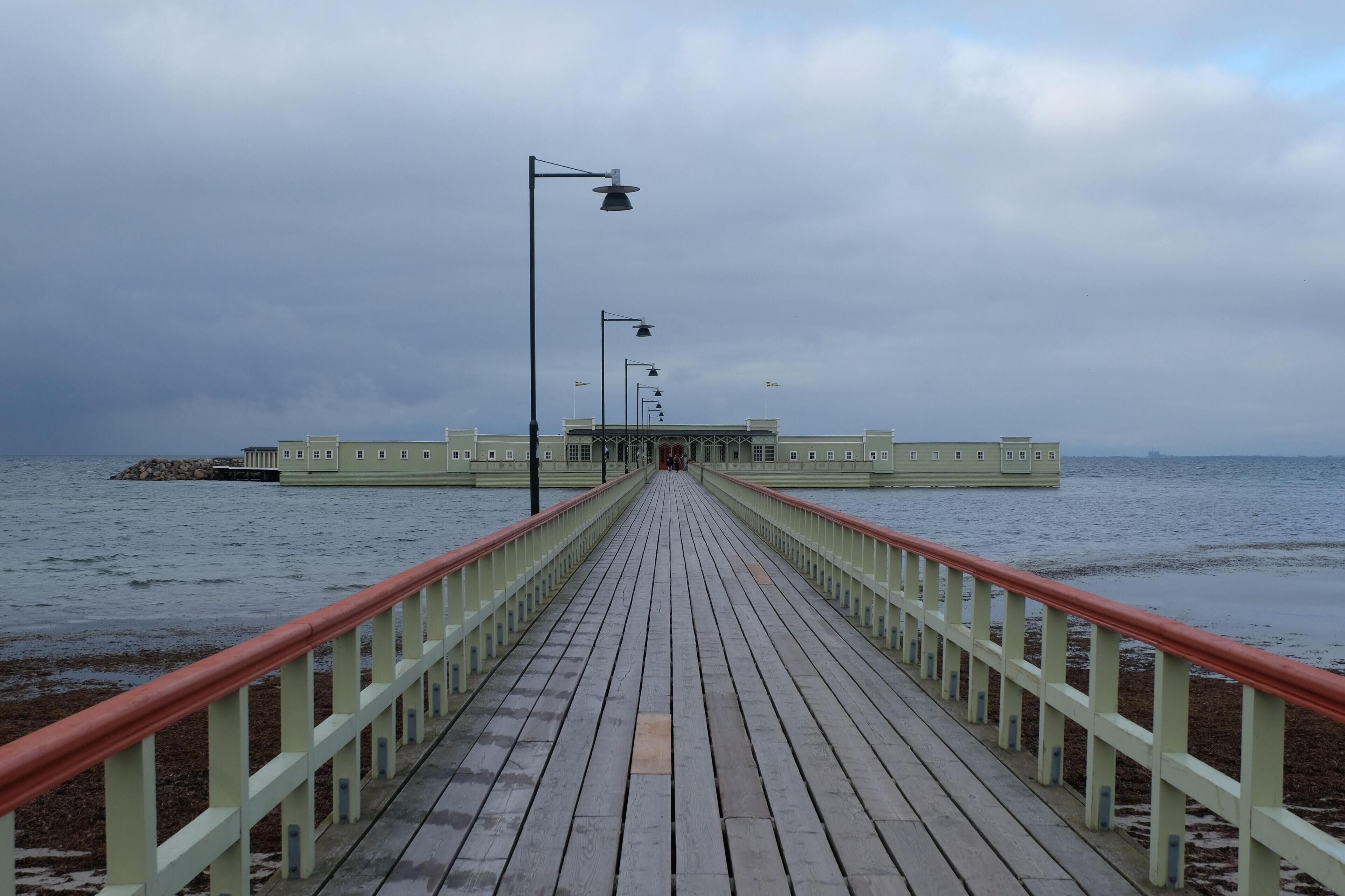 A wooden pier leading to a building with a light green facade, extending over a body of water.
